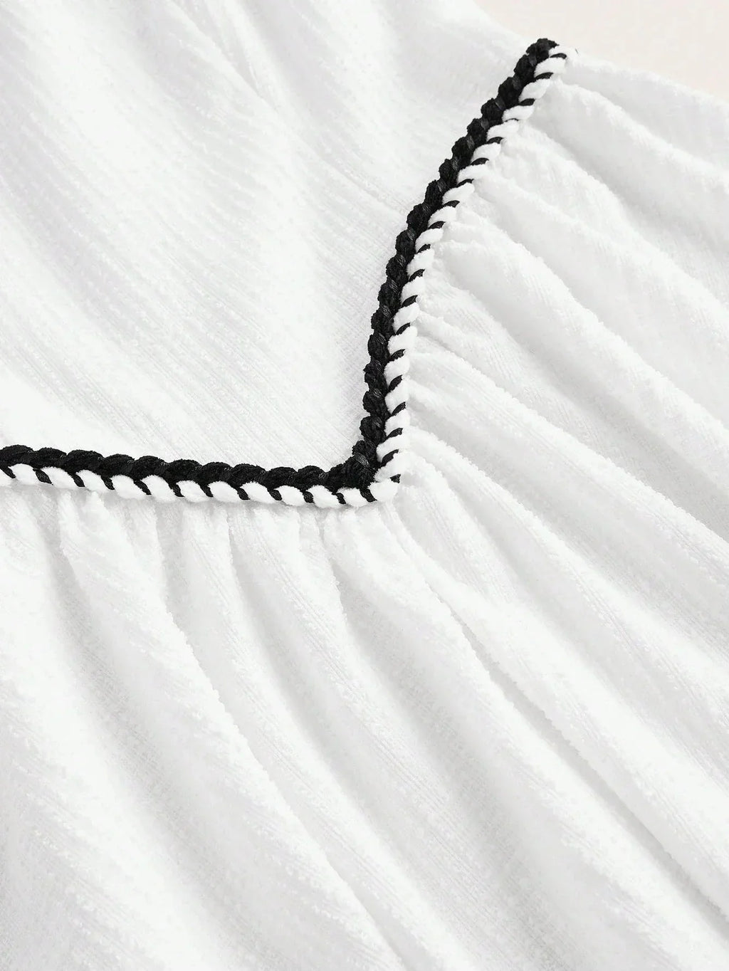 This charming white dress features delicate textured fabric with a playful black braided trim along the neckline, adding a.
