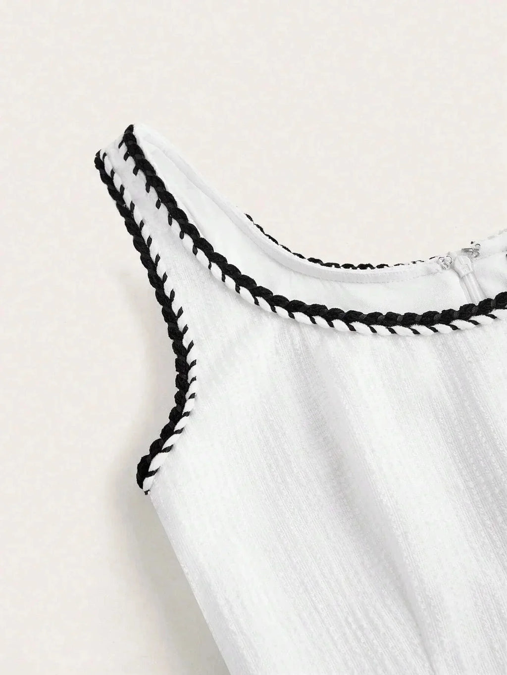 This elegant white dress features a flattering sleeveless design with a delicate black braided trim around the neckline,.