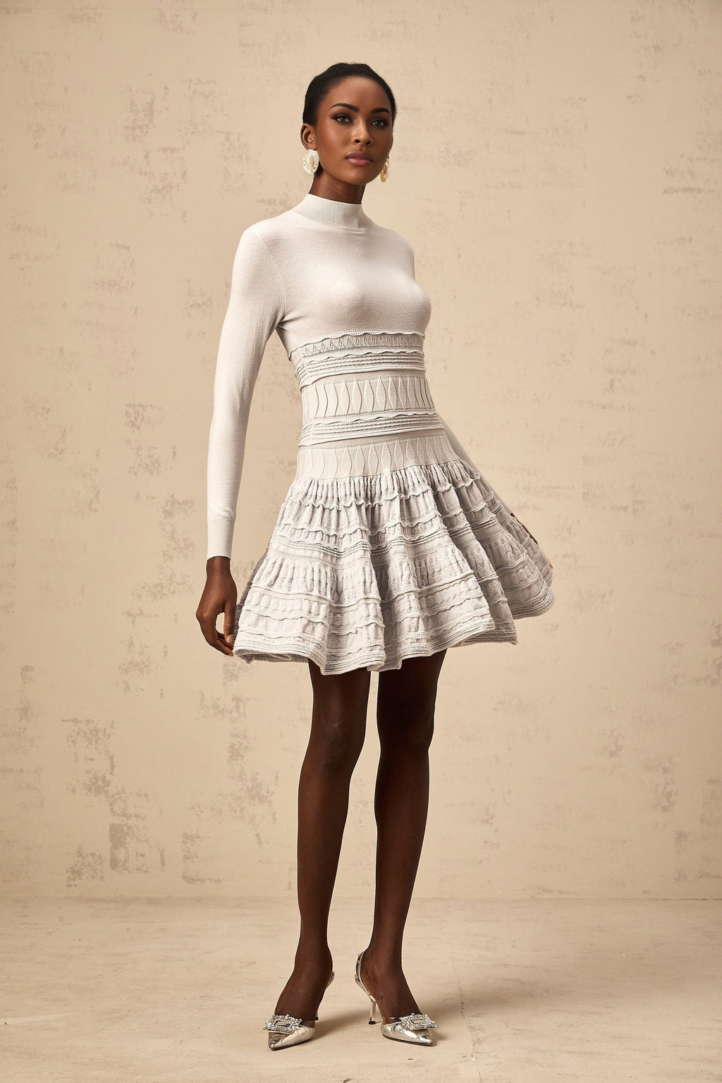 Woman wearing a short grey dress with ruffled details and ribbed texture standing against plain background